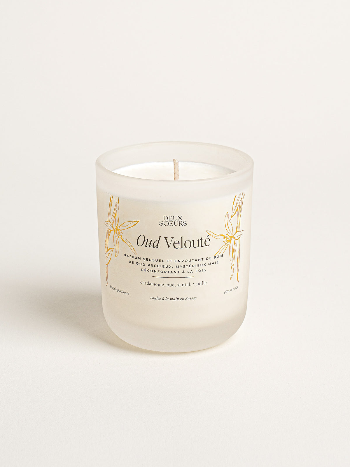 A frosted glass jar candle with white wax and a single wick, featuring gold botanical illustrations and French text on the label. The brand 'DEUX SOLEILS' is visible at the top of the label. The name of the fragrance is Oud Velouté.