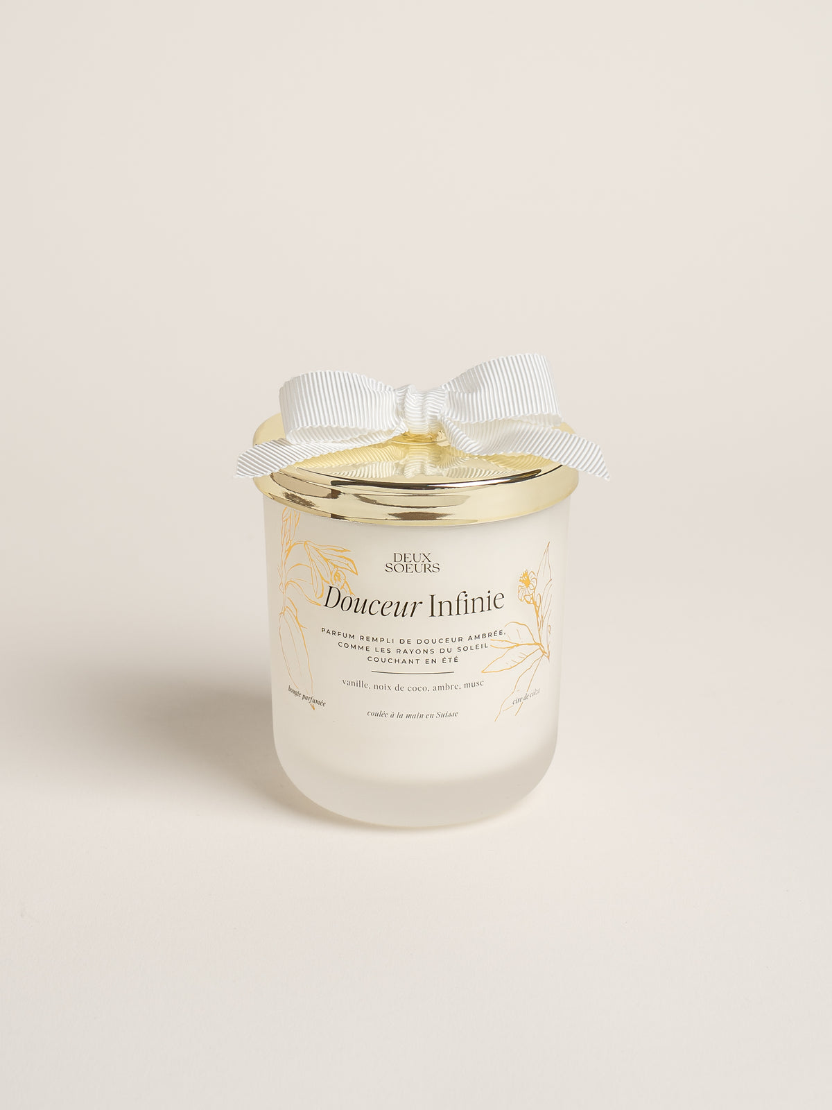 A frosted glass jar candle with a gold metal lid, decorated with a white satin bow. The label reads 'Douceur Infinitie' and features gold botanical illustrations, set against a solid off-white background.