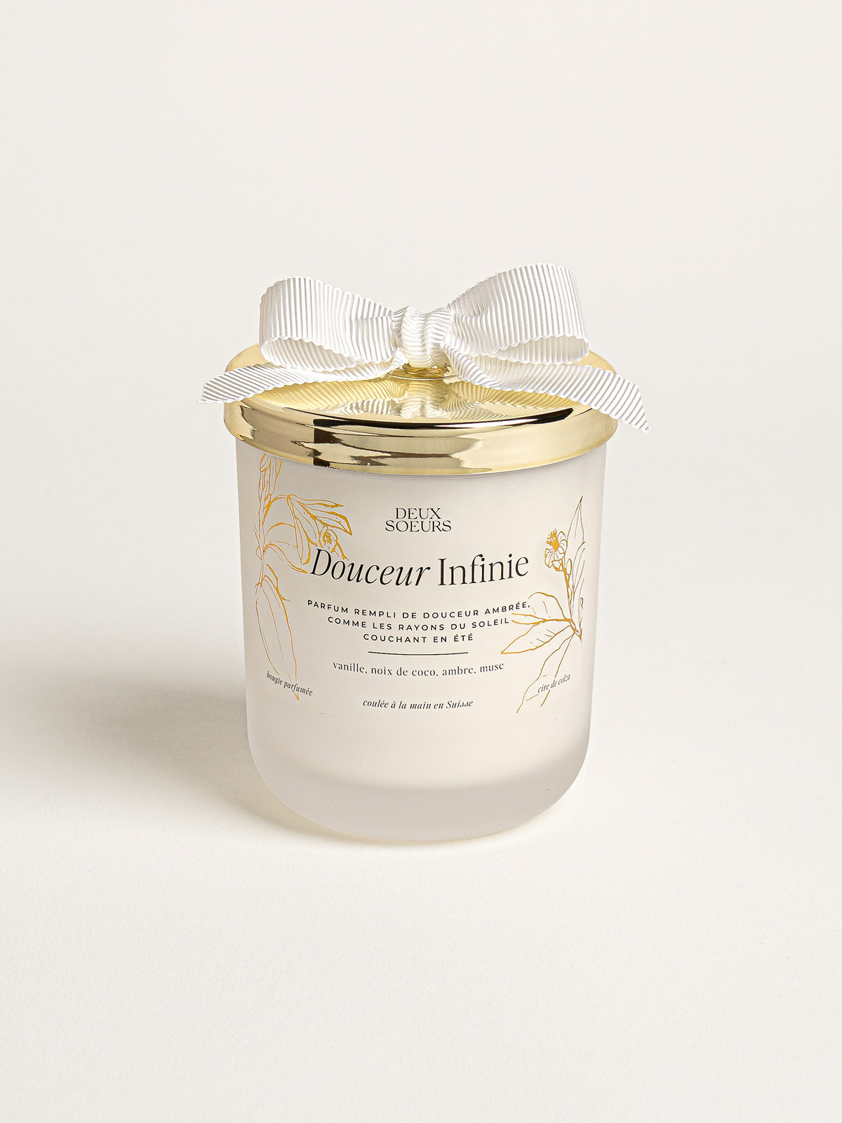 A frosted glass jar candle with a gold metal lid, decorated with a white satin ribbon bow. The label reads 'Douceur Infinie' and 'Deux Soeurs'.